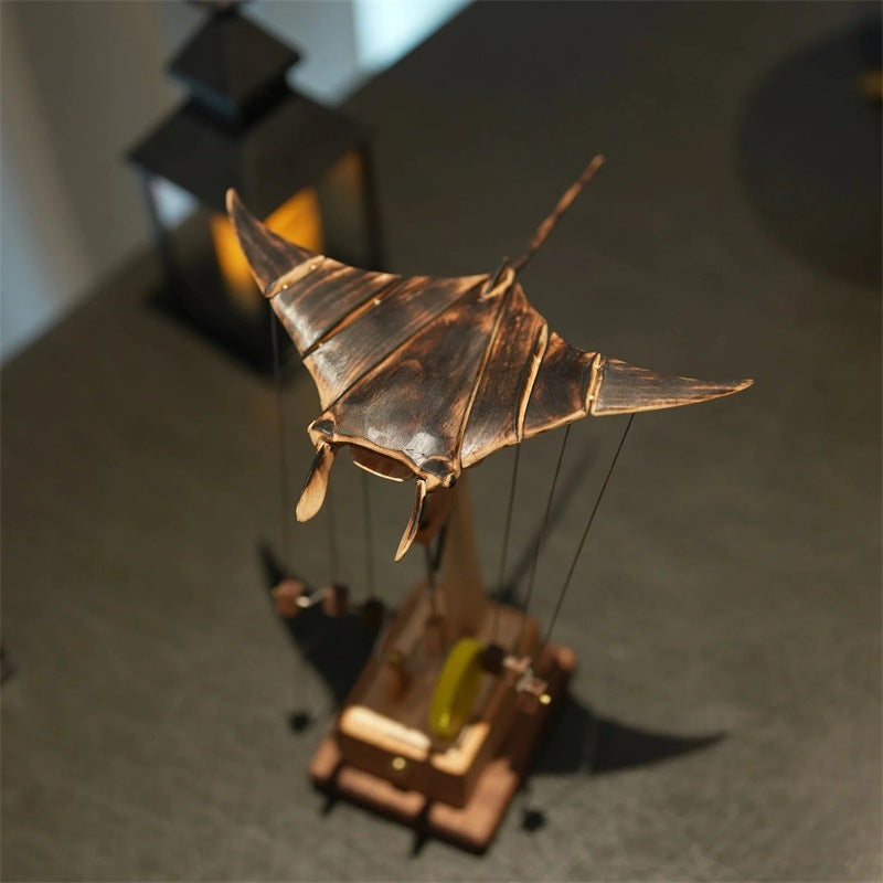 A wooden manta ray puppet toy on a wooden stand, photographed on a dark tabletop with a blurred lantern in the background. The TimberTide logo is visible in the bottom right corner.