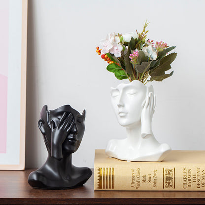 Decorative sculpture of a face with a hand covering one eye, next to a white vase with flowers on a book.
