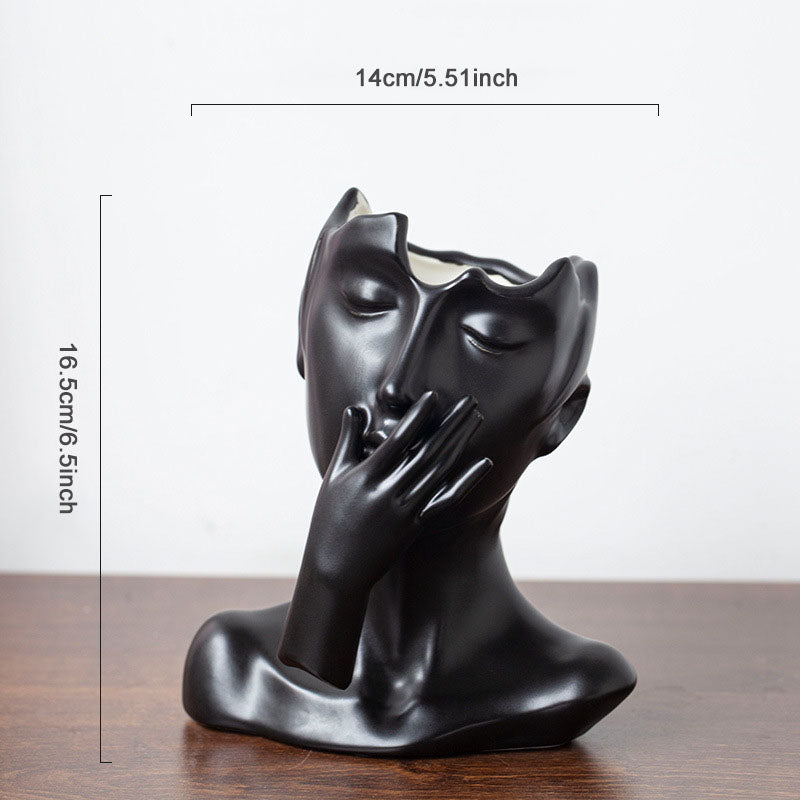Black decorative sculpture of a face with a hand over one eye on a wooden surface, with measurements indicated.